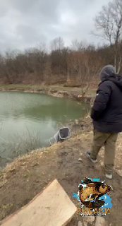 Рыболов идёт по краю водоёма в тёплой одежде; спокойная поверхность пруда, прибрежные кусты и логотип канала на кадре.