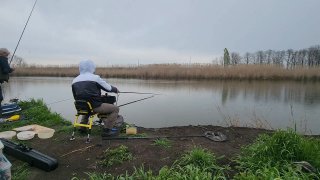 Ещё один кадр с рыбаками на берегу: сидящие участники в дождевиках, удилища в рабочем положении и мокрая почва у воды при сером небе.