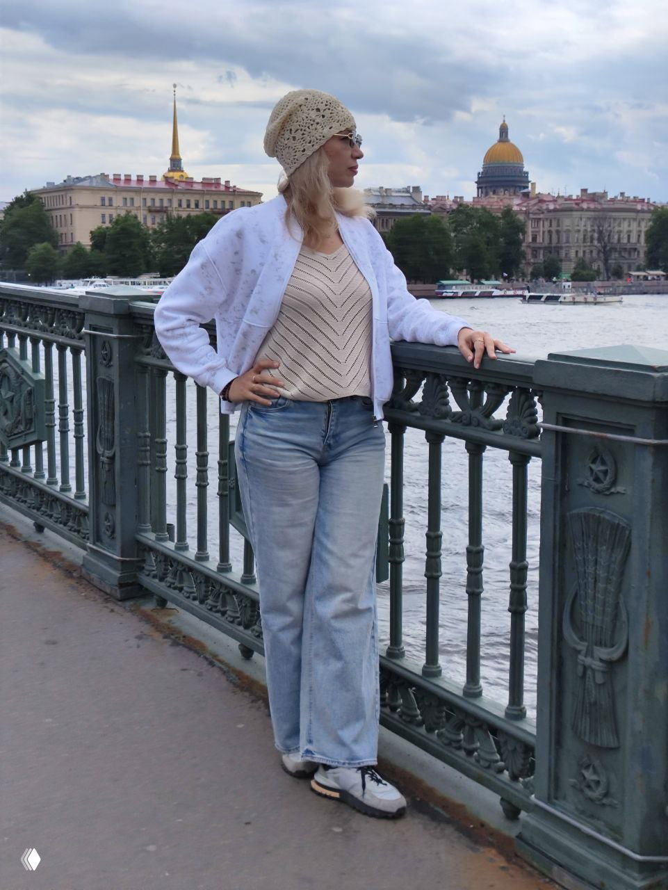 Женщина в лёгкой куртке стоит у перил на набережной Невы; вдали виднеется Исаакиевский собор и городской пейзаж.