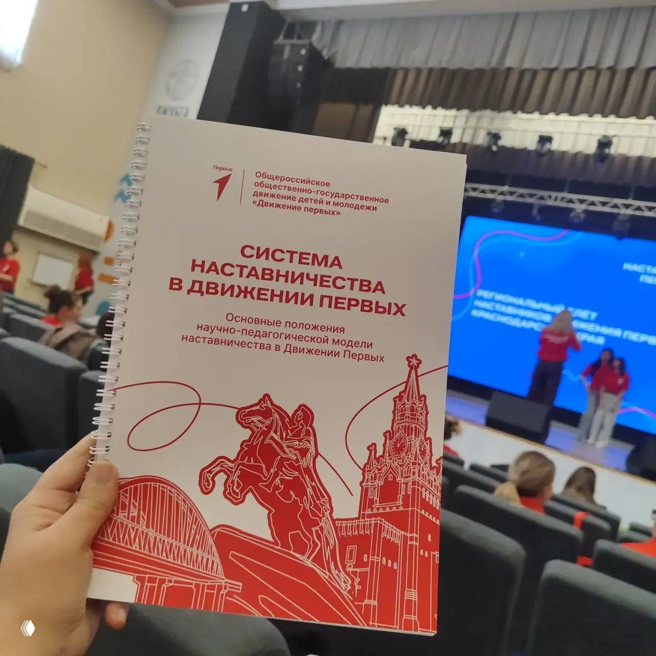 Рука держит брошюру «Система наставничества в Движении Первых» на фоне сцены и зала; видны заголовок и иллюстрации.