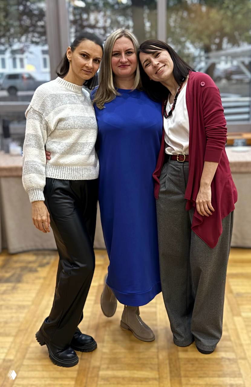 Три женщины улыбаются и обнимаются у окна на финальном событии проекта «Семейный остров» в Туапсе, групповое фото.