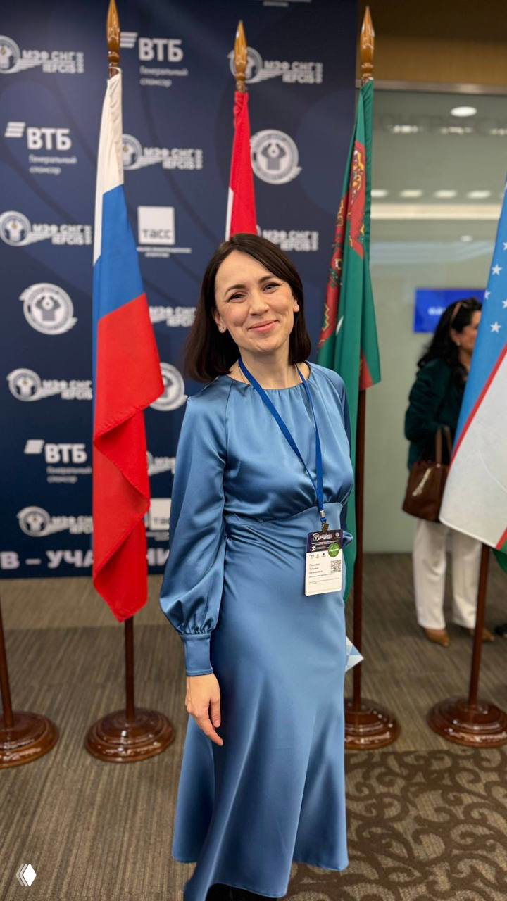 Автор поста в синем платье с бейджем участника на фоне флагов государств‑участников СНГ в конференц-зале во время международного экономического форума