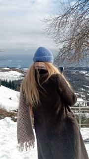 Женщина в голубой шапке и тёплом пальто стоит у перил смотровой площадки около Мураззано и смотрит на заснеженные лавандовые поля и холмы.