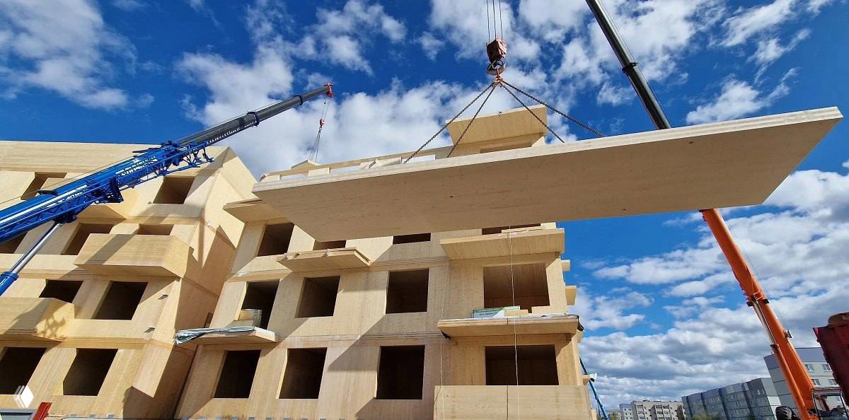 Кран поднимает крупную сборную деревянную панель на стройплощадке многоквартирного дома из клеёного бруса под частично облачным небом.