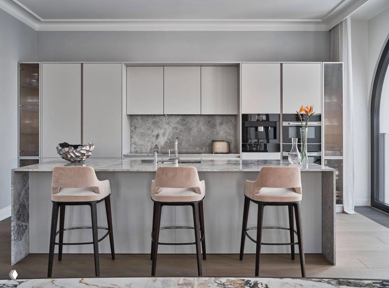 Кухня с островом и тремя барными стульями, светлая мебель и мраморная рабочая поверхность, часть пространства открытой планировки.