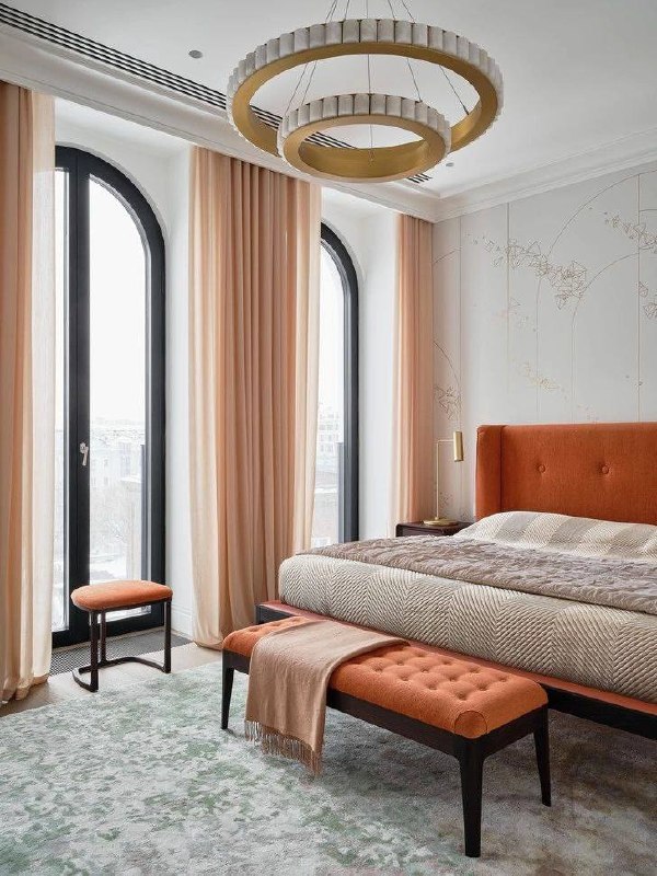 Спальня с высокими арочными окнами, оранжево‑терракотовая обивка кровати и нежные шторы — тёплая и уютная атмосфера комнаты.
