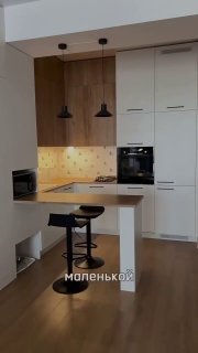 Небольшая функциональная кухня с барной стойкой, двумя подвесными светильниками, встроенной духовкой и белыми шкафами, деревянный пол и акценты.