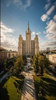 Фасад сталинской высотки в центре города, неоклассическое здание с башней на фоне синего неба и аллеей на переднем плане