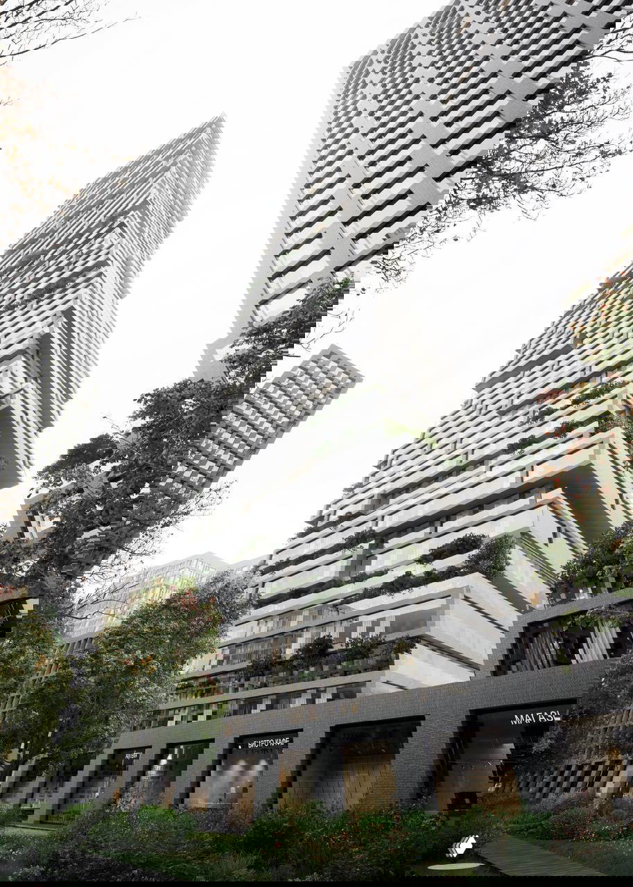 Крупный план высотного объёма с выраженным массингом и многоуровневыми террасами, пространство у подножия с магазинами и деревьями.