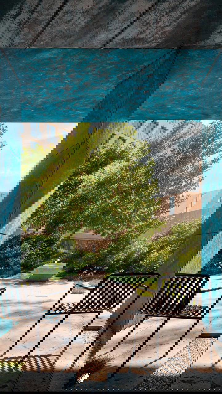 Кадр с выходом во внутренний двор: синие облицовочные панели обрамляют вид на зелёный двор с деревьями и свободными зонами отдыха, видны металлические кресла на переднем плане.