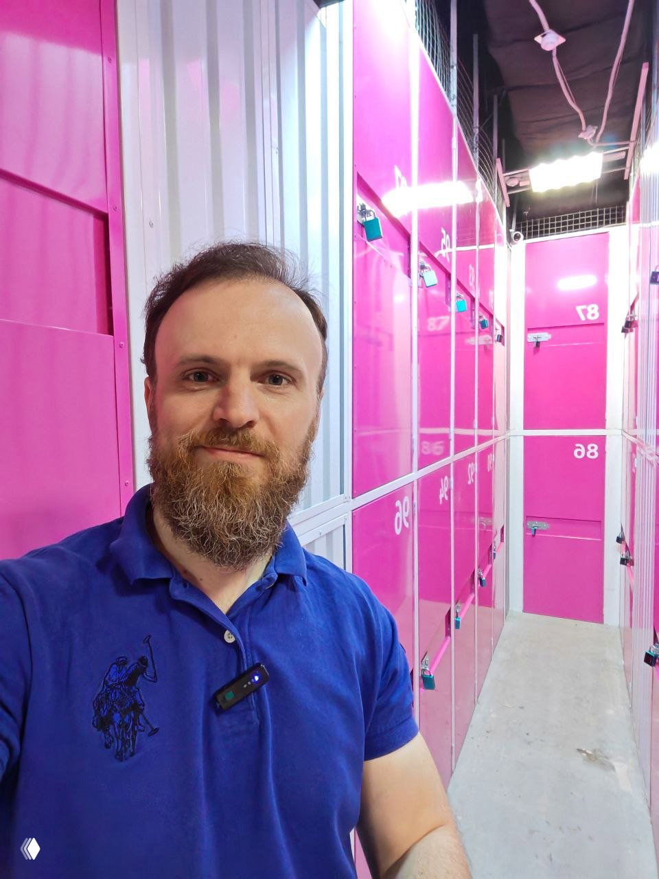 Мужчина с бородой делает селфи в коридоре ярко-розовых ячеек self-storage; видны металлические дверцы, замки и номера.