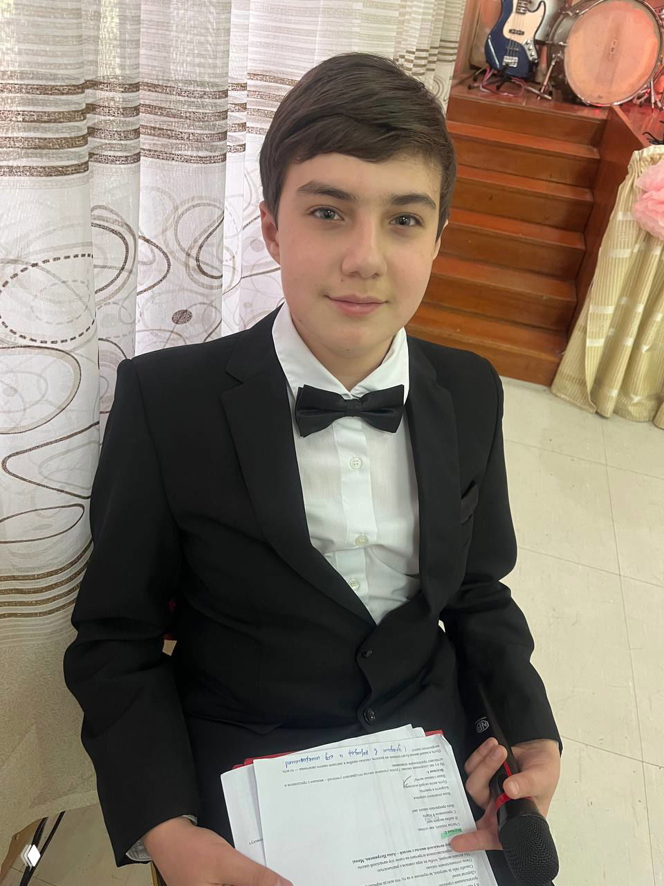 Крупный план подростка в белой рубашке и бабочке, в руках блокнот с текстом программы и микрофон — подготовка к роли ведущего на концерте.