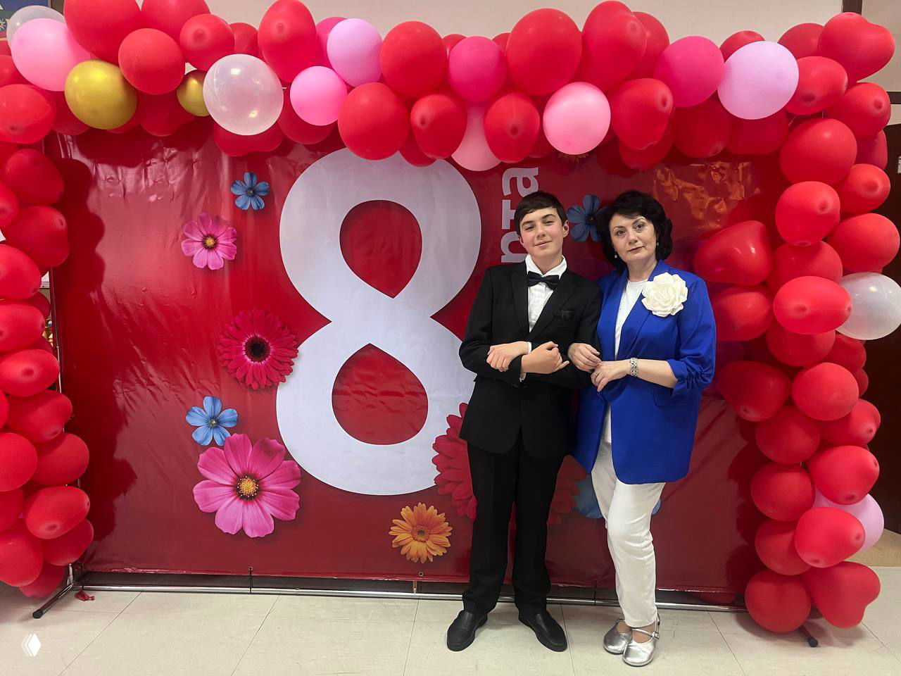Подросток в смокинге рядом с женщиной на фоне арки из красных и розовых шаров и большой цифры 8 — общий кадр после школьного мероприятия.