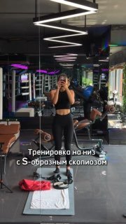 Женщина в спортзале снимает себя на телефон на фоне зеркал и тренажёров; текст на фото: «Тренировка на-низ с S-образным сколиозом».