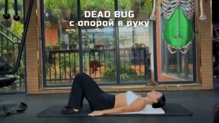 Женщина на коврике в зале демонстрирует вариацию упражнения Dead Bug с опорой на руку; видно положение корпуса и согнутые колени для контроля поясницы.