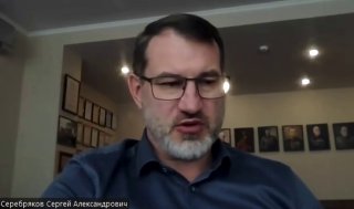 Кадр из видео: мужчина в очках ведёт разговор в формате видеозаписи, на заднем плане видна стена с портретами и потолочные светильники.