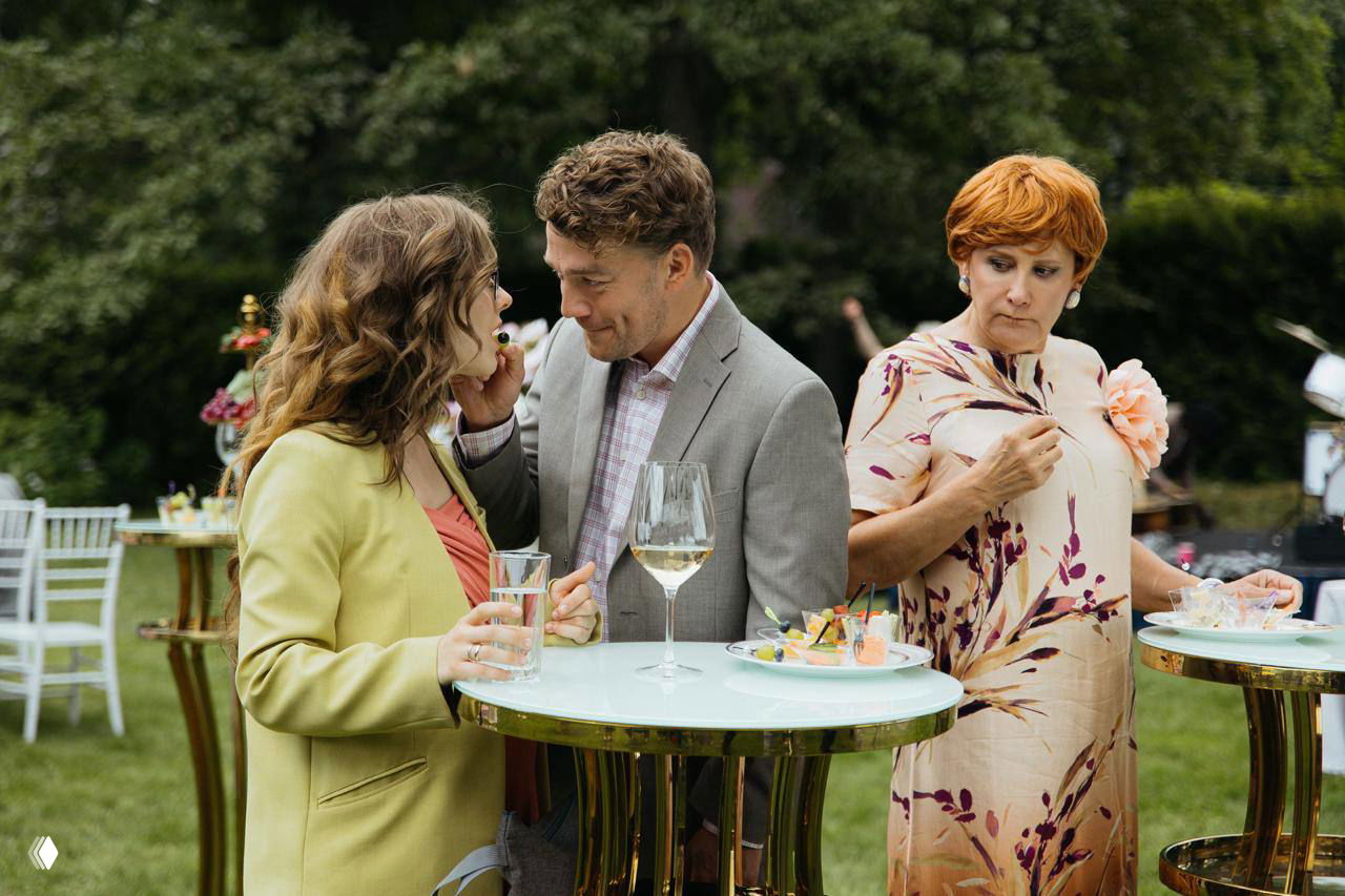 Открытая сцена на крытой веранде: трое персонажей общаются за высоким столиком, на переднем плане бокал вина и закуски — фрагмент праздничной встречи в фильме.