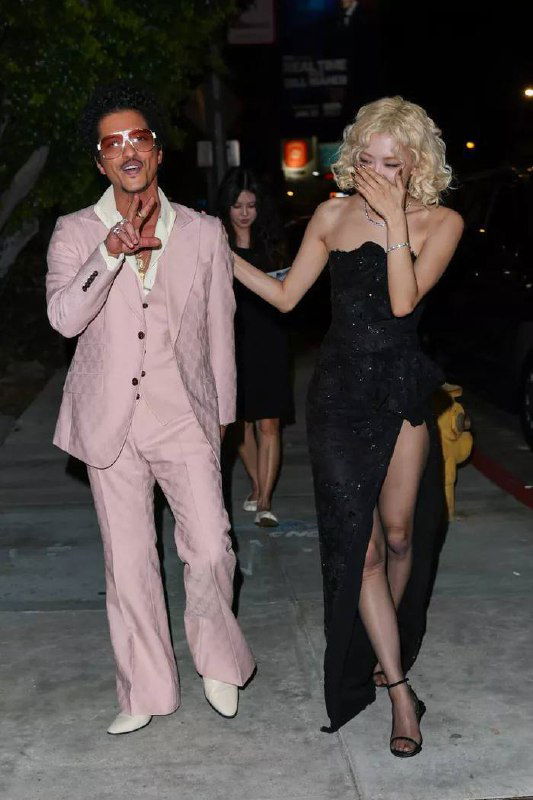 Пара идёт по ночному тротуару: мужчина в розовом костюме машет рукой, женщина в чёрном платье прикрывает рот и смеётся