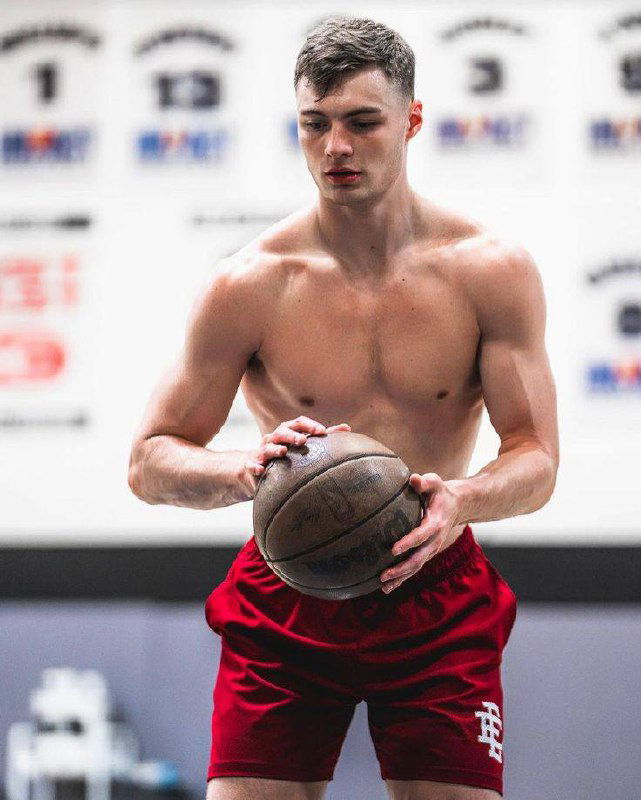 Мужчина‑спортсмен без майки держит баскетбольный мяч в зале; кадр подчёркивает мускулатуру, концентрацию и тренировочный процесс