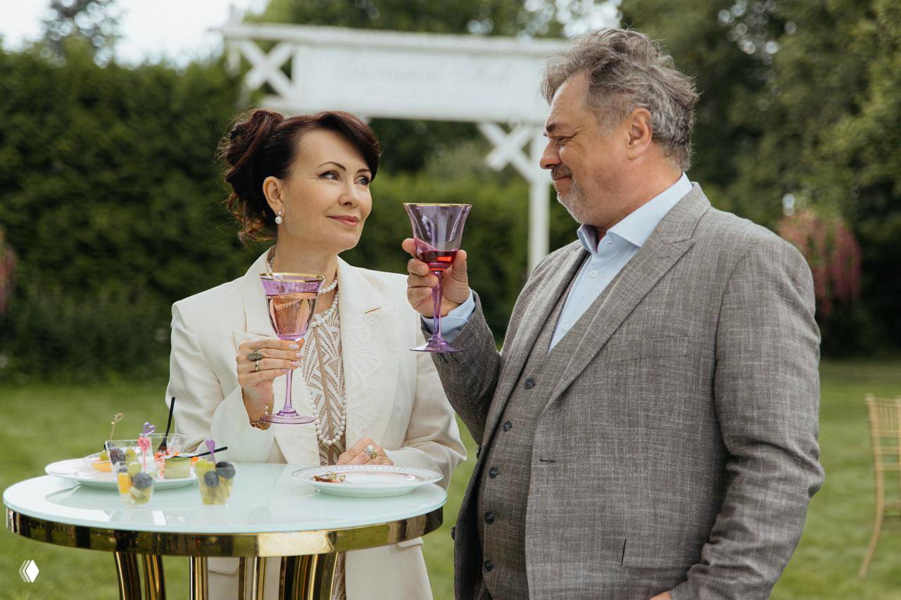 Пара взрослых персонажей поднимает бокалы на садовой вечеринке: женщина в светлом костюме и мужчина в сером пиджаке в тёплый момент диалога.