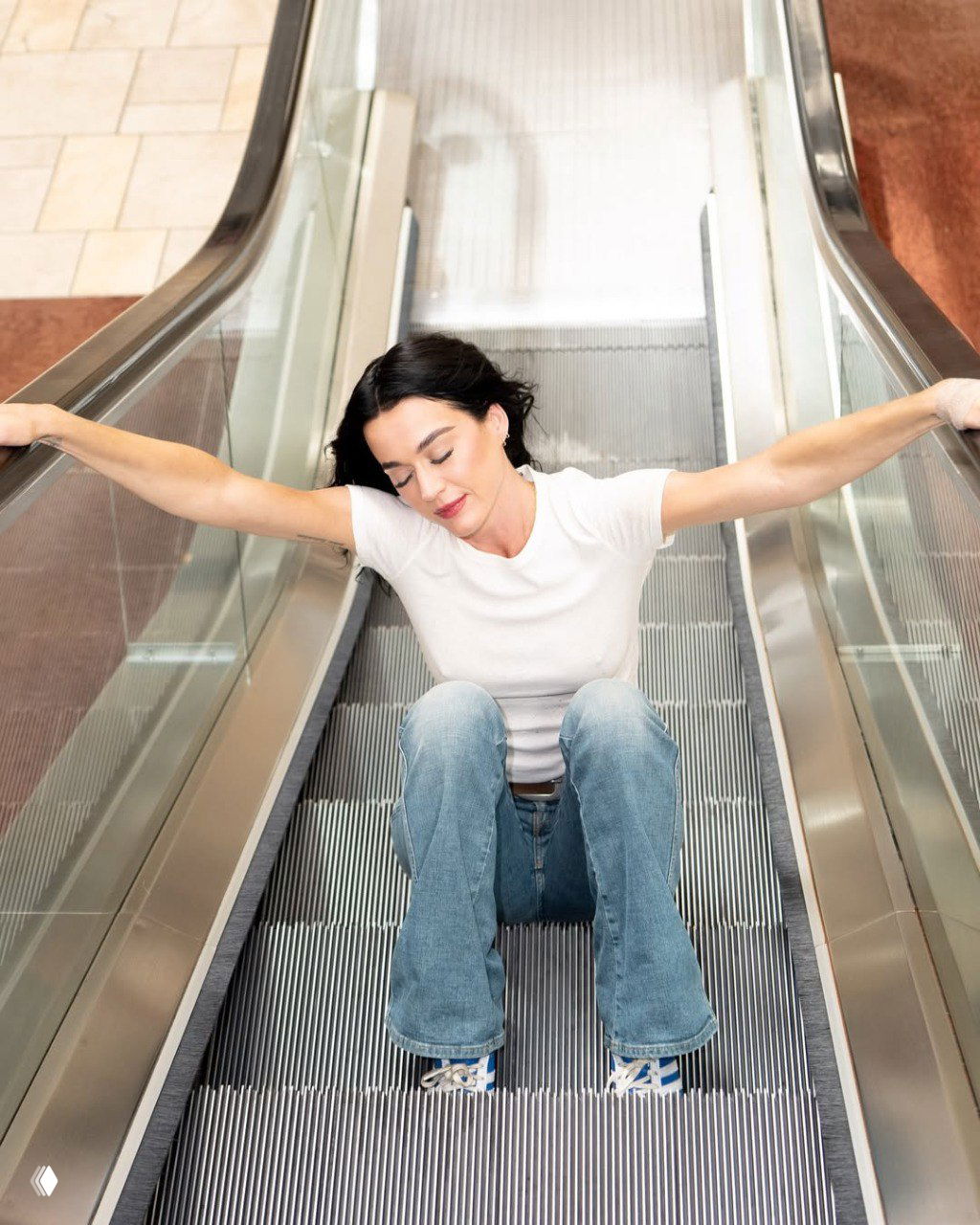 Женщина сидит на ступенях эскалатора, держась за поручни по обе стороны; кадр показывает позу и интерьер торгового центра.