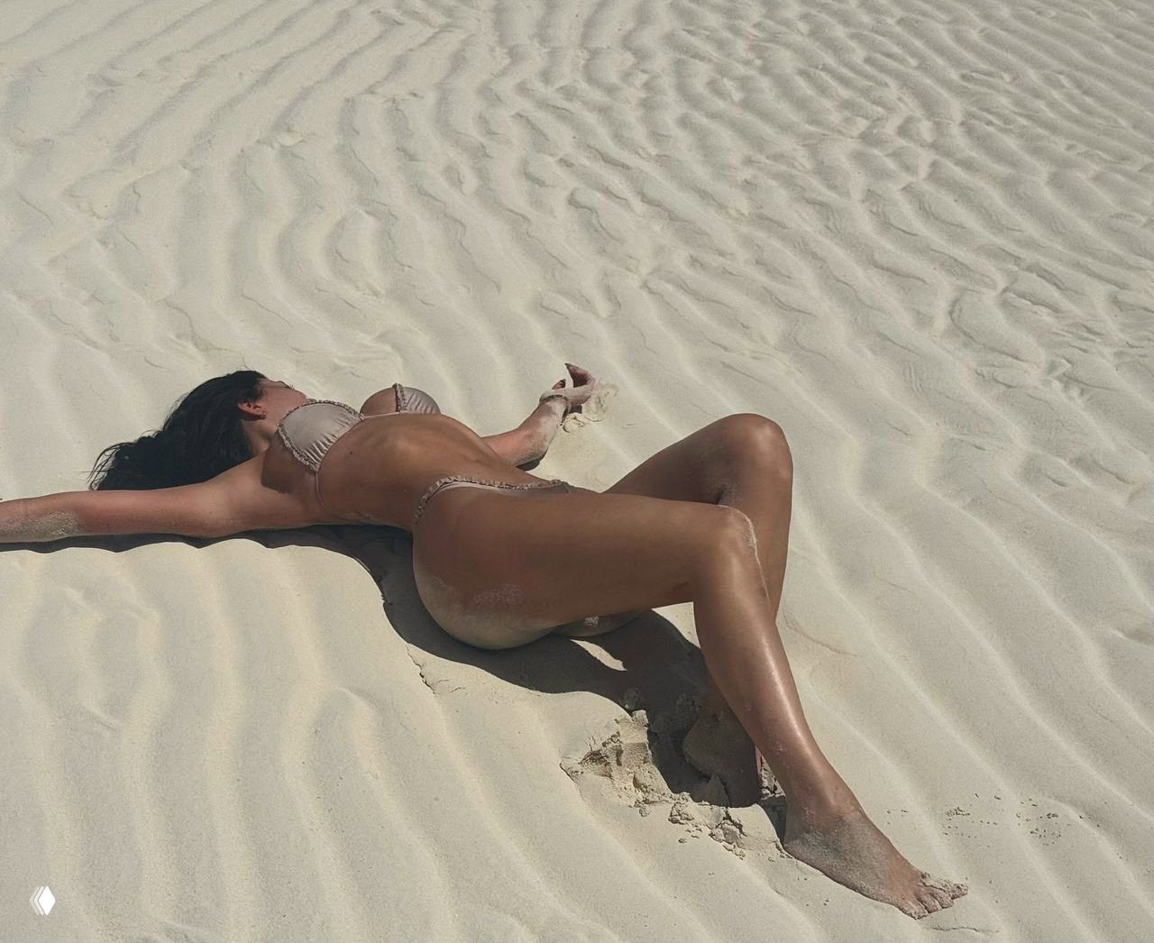 Женщина в бикини лежит на рифлёном белом песке островного пляжа; видны следы на песке, тени и текстура поверхности вокруг.