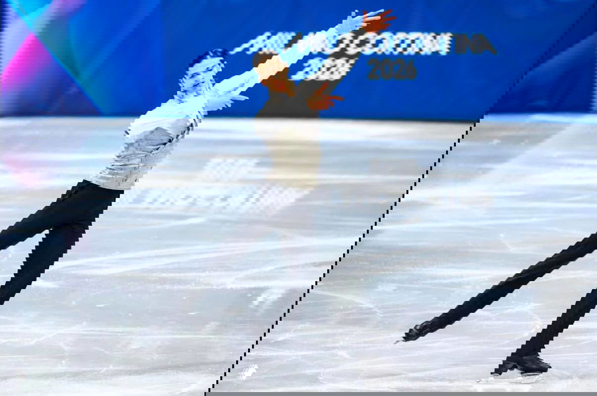 Фигурист в костюме исполняет программу на ледовой арене, видно поверхность льда и баннер соревнований на заднем плане