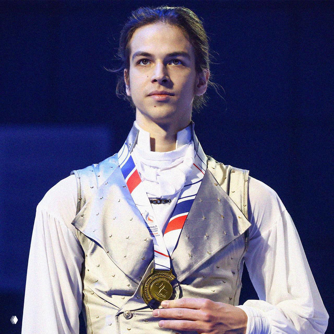 Портрет фигуриста в светлом сценическом костюме с медалью на груди, крупный план лица на фоне тёмной сцены.