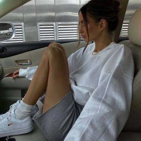 Девушка в салоне машины в светлом свитере и спортивных шортах прислонилась к двери — повседневный минималистичный образ. Фото в стиле эстетики: нейтральная палитра и мягкое освещение.