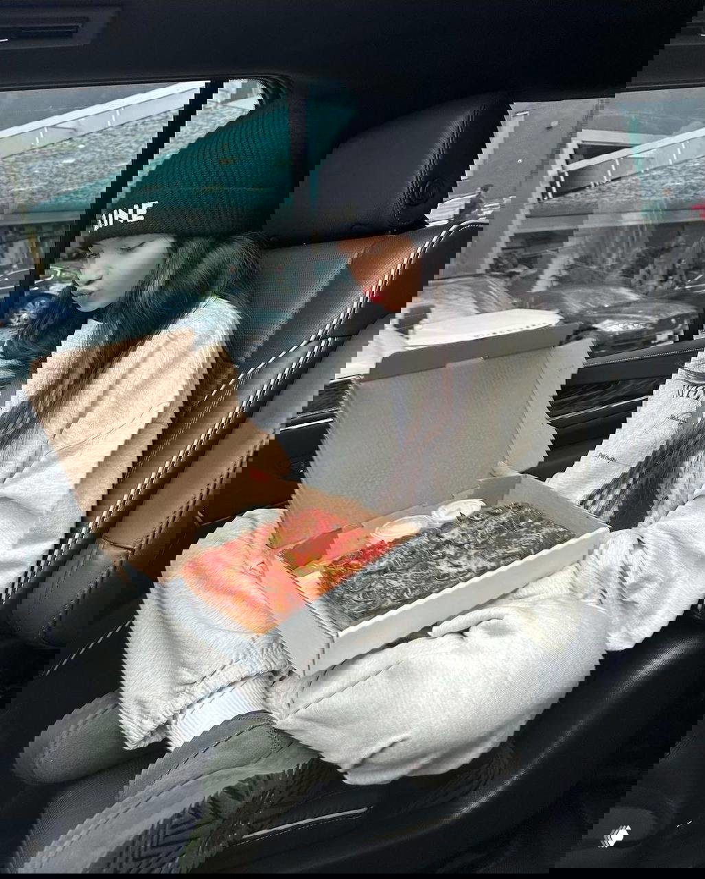 Лиса из BLACKPINK смотрит на открытый контейнер с пиццей в машине; видно пепперони и приправы, рядом соусы.