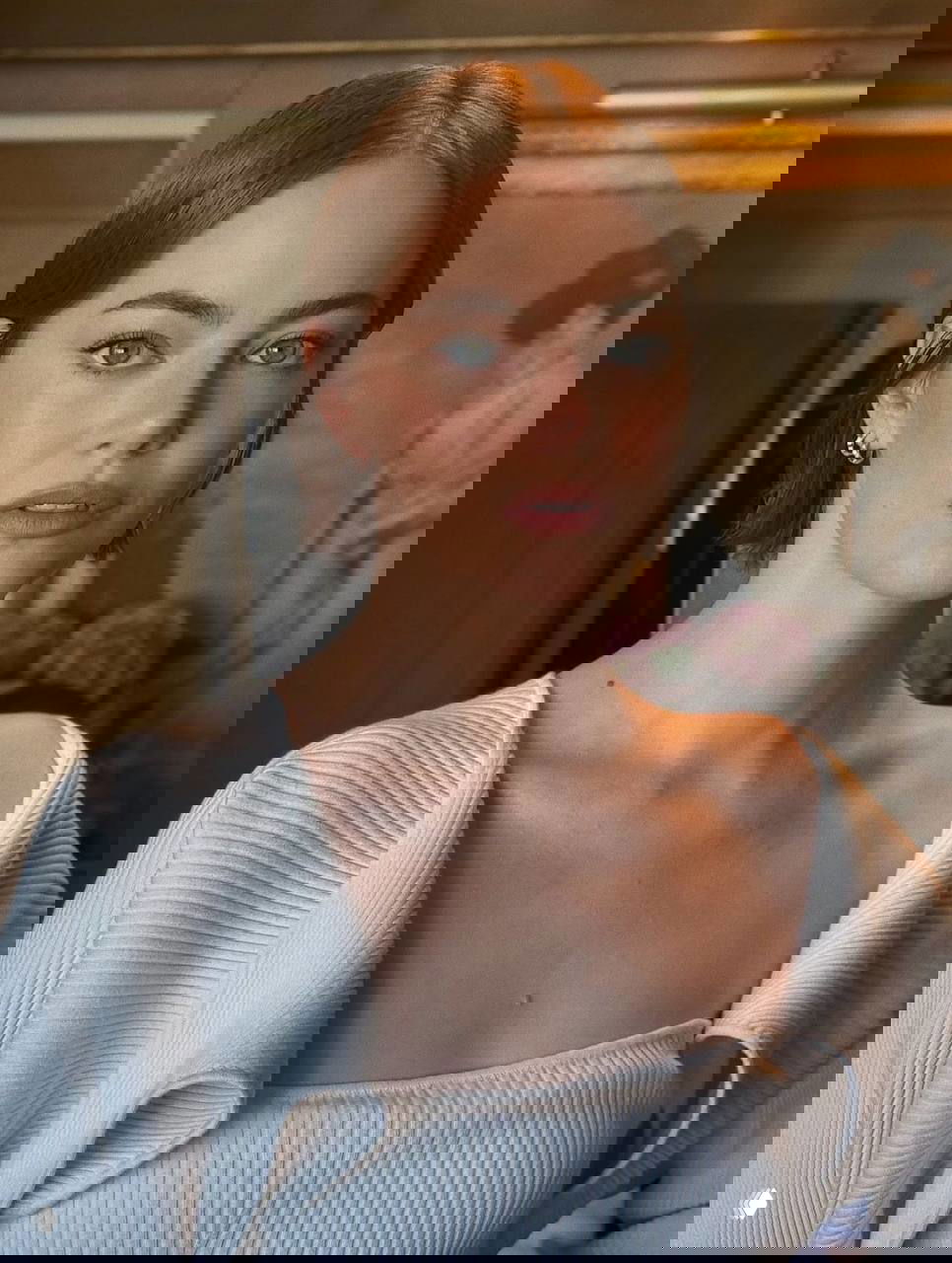 Портрет Эммы Стоун в светлом трикотажном топе, естественный макияж и открытые плечи на фоне комнаты с цветами и картиной.