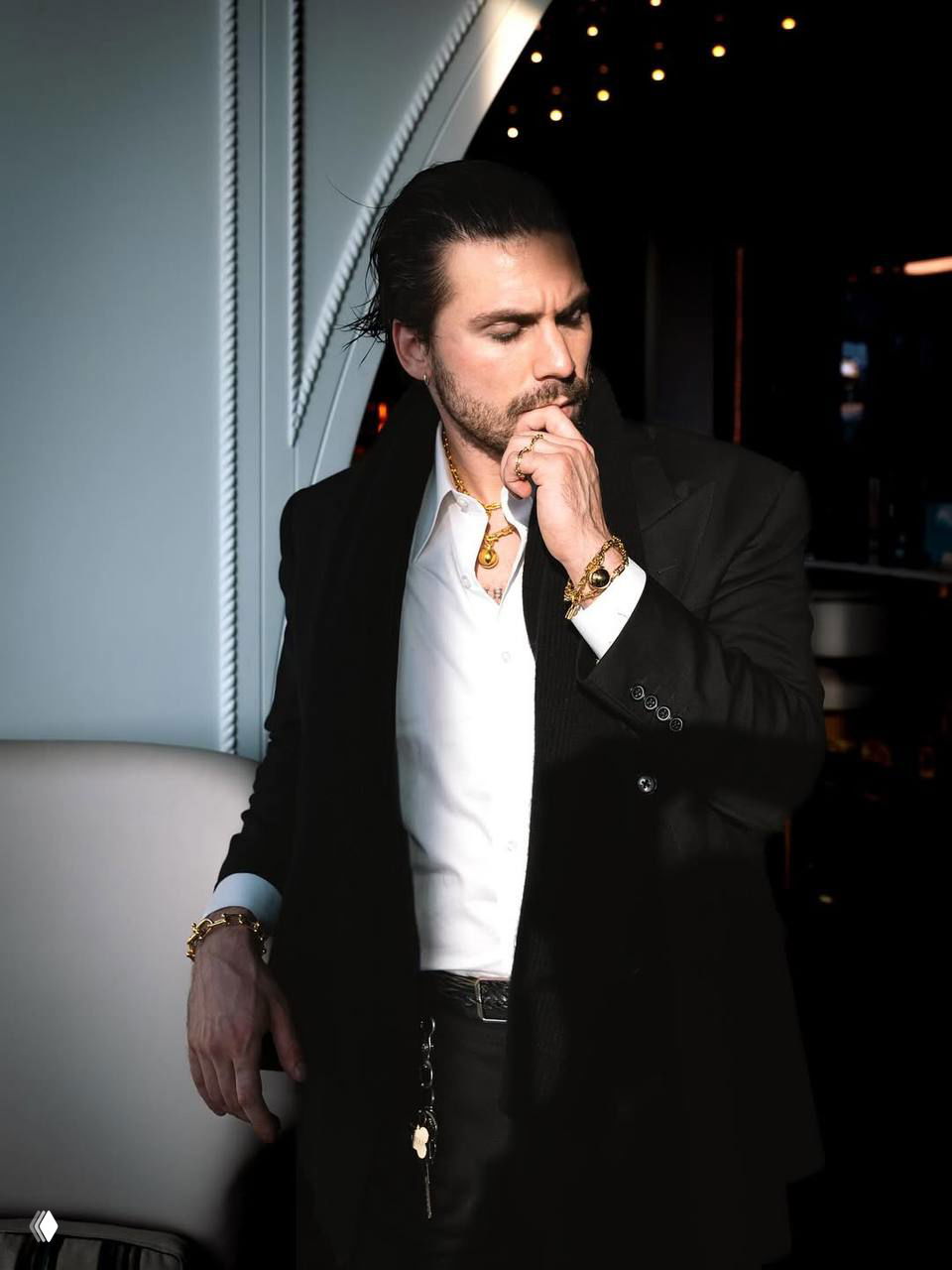 Мужской портрет в полный рост: тёмный пиджак, белая рубашка и золотые украшения на фоне светлого декора и мягкого боке.