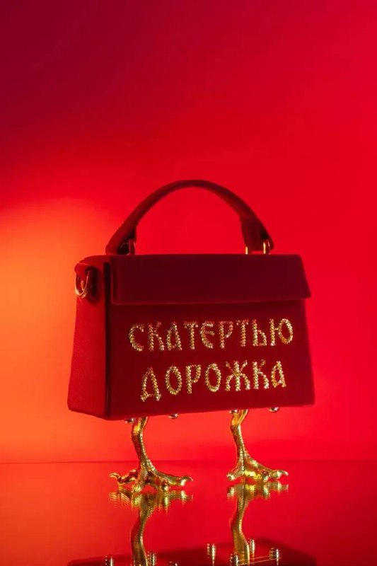 Яркая студийная фотография: красная дизайнерская сумка на золотых куриных лапках на контрастном фоне, с вышитой надписью «СКАТЕРТЬЮ ДОРОЖКА».