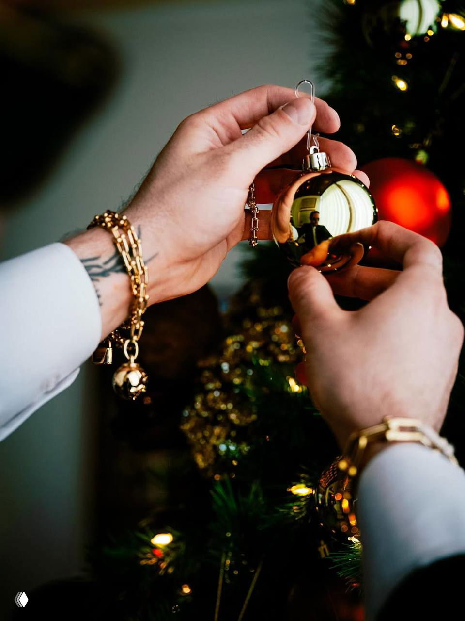 Крупный план рук мужчины с татуировкой и золотыми браслетами, он держит блестящий ёлочный шар с заметным отражением внутри.