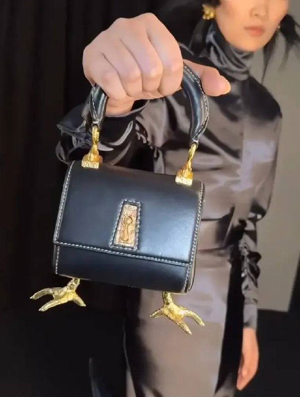 Черная мини-сумка на золотых куриных лапках держится рукой модели в блестящем атласном платье; видно фокус на фурнитуре, швах и декоративных лапках.