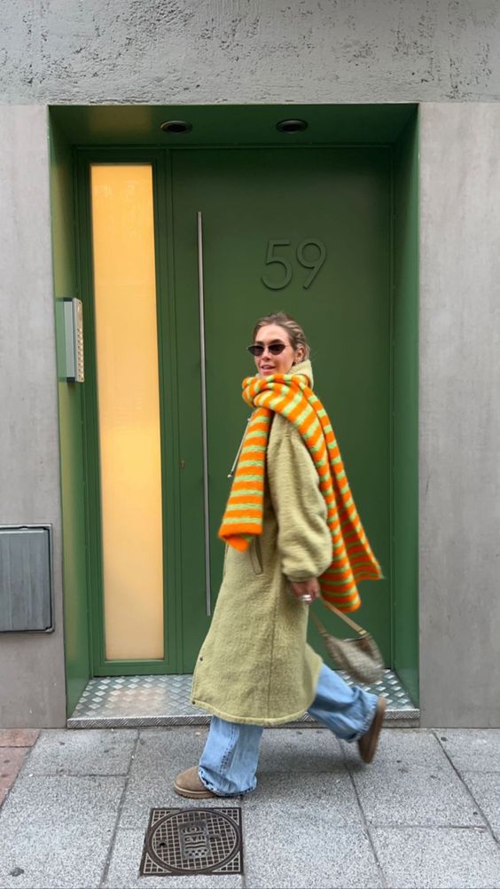 Женщина идёт по улице в полосатом оранжево-зелёном шарфе большого размера и светлом пальто — контрастные оттенки в городском образе.