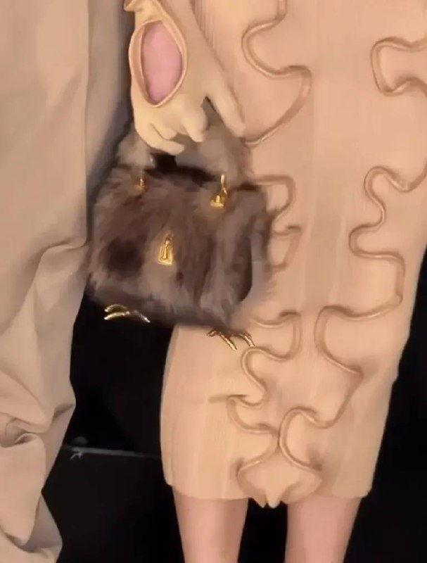 Меховая сумка на золотых куриных лапках висит у руки модели в бежевом наряде; кадр подчёркивает текстуру меха и декоративные лапки как акцент коллекции.