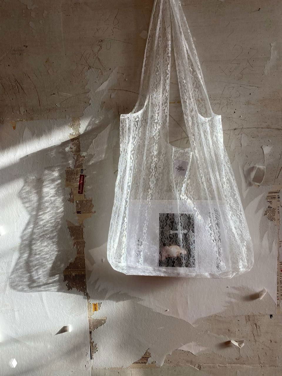 Прозрачная кружевная сумка на текстурной стене: лёгкая ткань, тени и художественный беспорядок на фоне подчёркивают фактуру кружева.