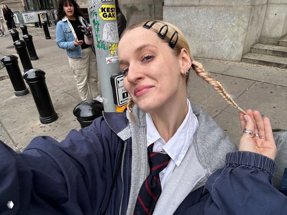 Крупный план женщины на улице: она делает селфи, вокруг видны столбики, прохожие и городская уличная среда в городской зоне.