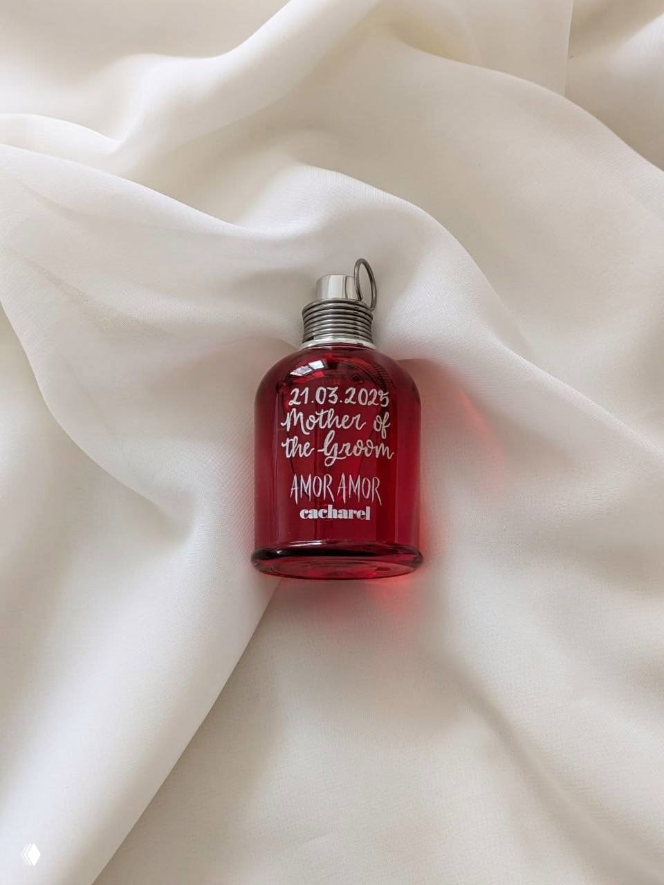 Мини‑флакон Cacharel Amor Amor тёмно‑красного цвета на мягкой белой ткани; видно металлическую крышку и декоративную надпись на бутылочке, стилистика пресс‑фото аромата.