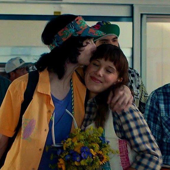 Съёмочный кадр: юноша целует девушку в щёку, они обнимаются на фоне людного пространства, в руках — букет цветов, тёплая сцена из эпизода.