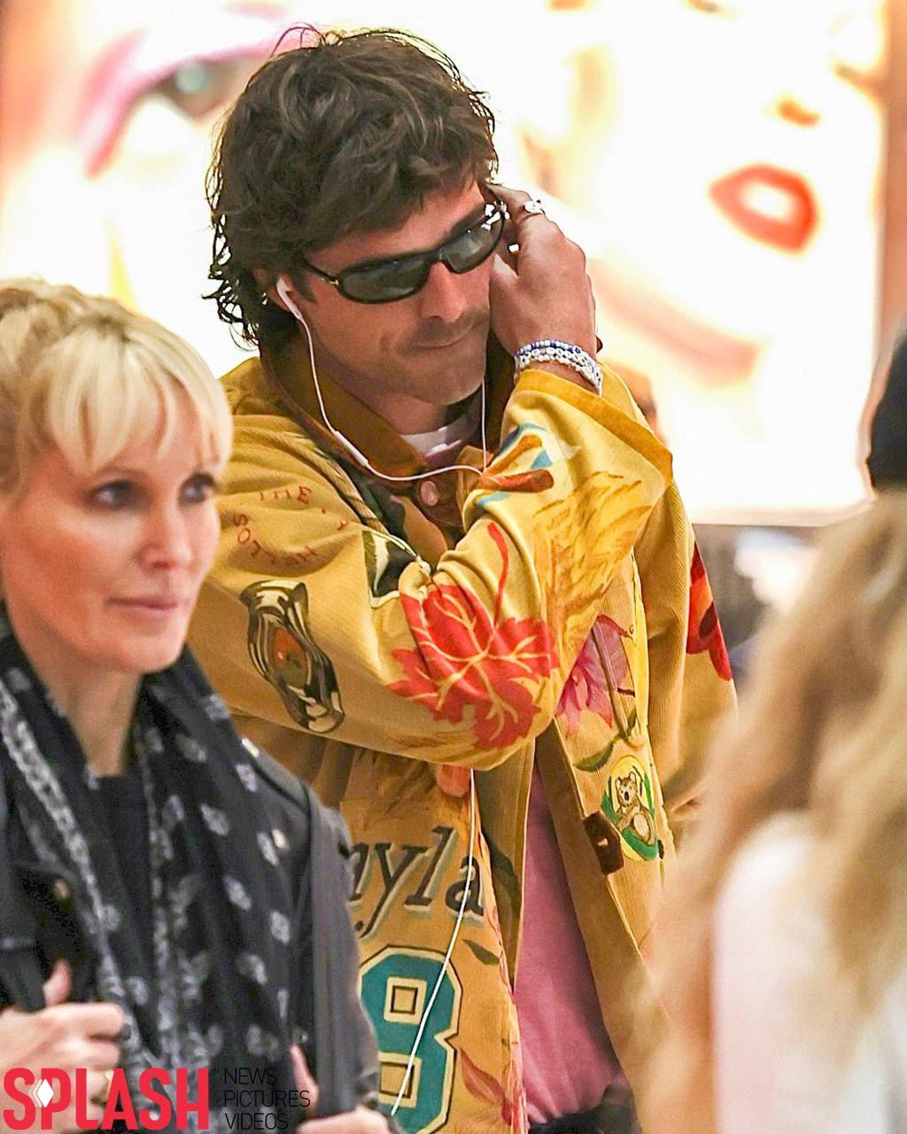 Крупный план мужчины в тёмных очках: в ушах видны белые проводные наушники, на руке браслеты и украшения, фон — интерьер терминала