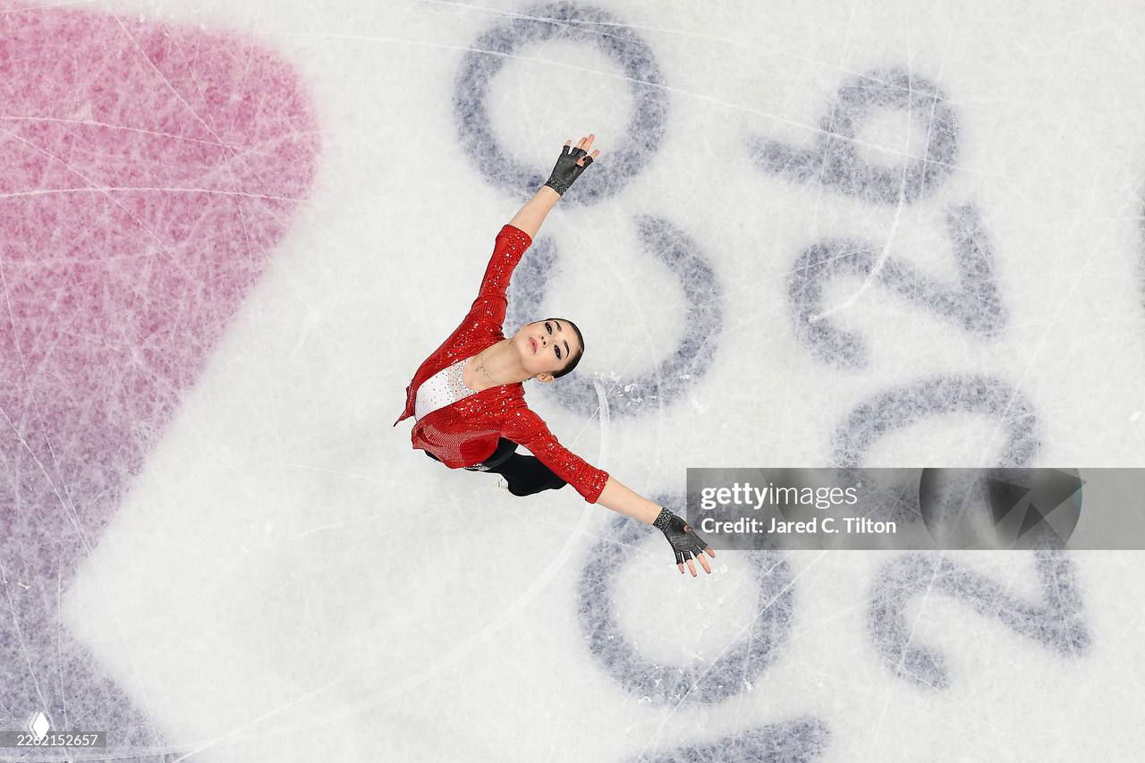 Аэрофотоснимок: Аделия Петросян в красном костюме исполняет элементы на льду рядом с символикой Олимпиады, видна структура замороженного покрытия и следы коньков.