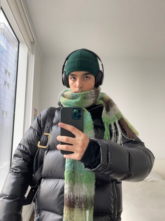 Селфи в зеркале: человек в чёрном пуховике и полосатом зелёно-бежевым шарфе с наушниками — уютный городской вариант для холодов.