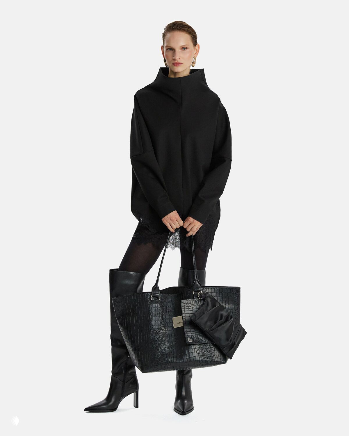 Модель в чёрной оверсайз тунике и высоких сапогах держит крупную кожаную сумку чёрного цвета — образ минималистичной городской элегантности LIZURA.
