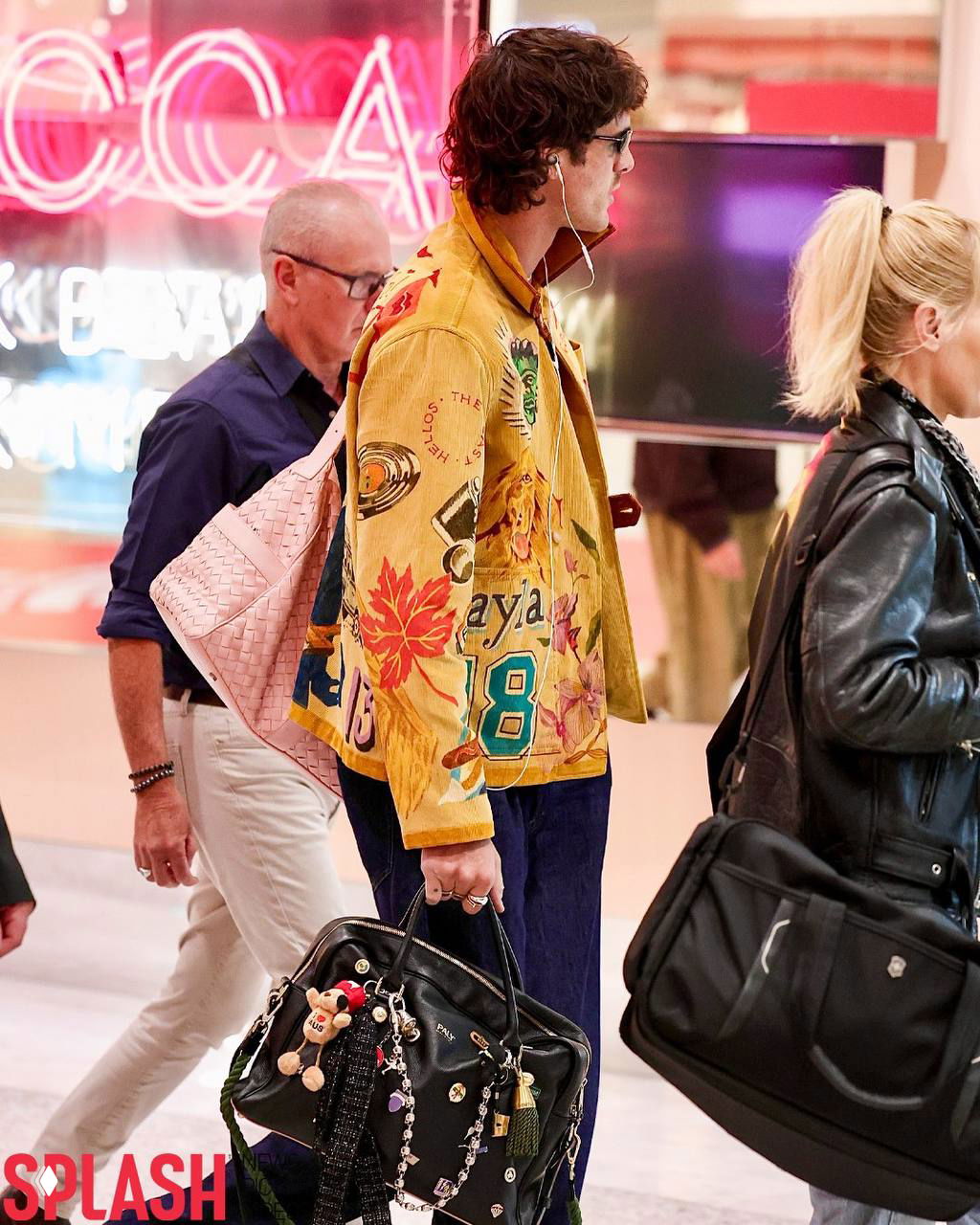 Человек в жёлтой вышитой куртке и тёмных штанах проходит по очереди в аэропорту, в ушах белые проводные наушники и большая чёрная сумка в руке