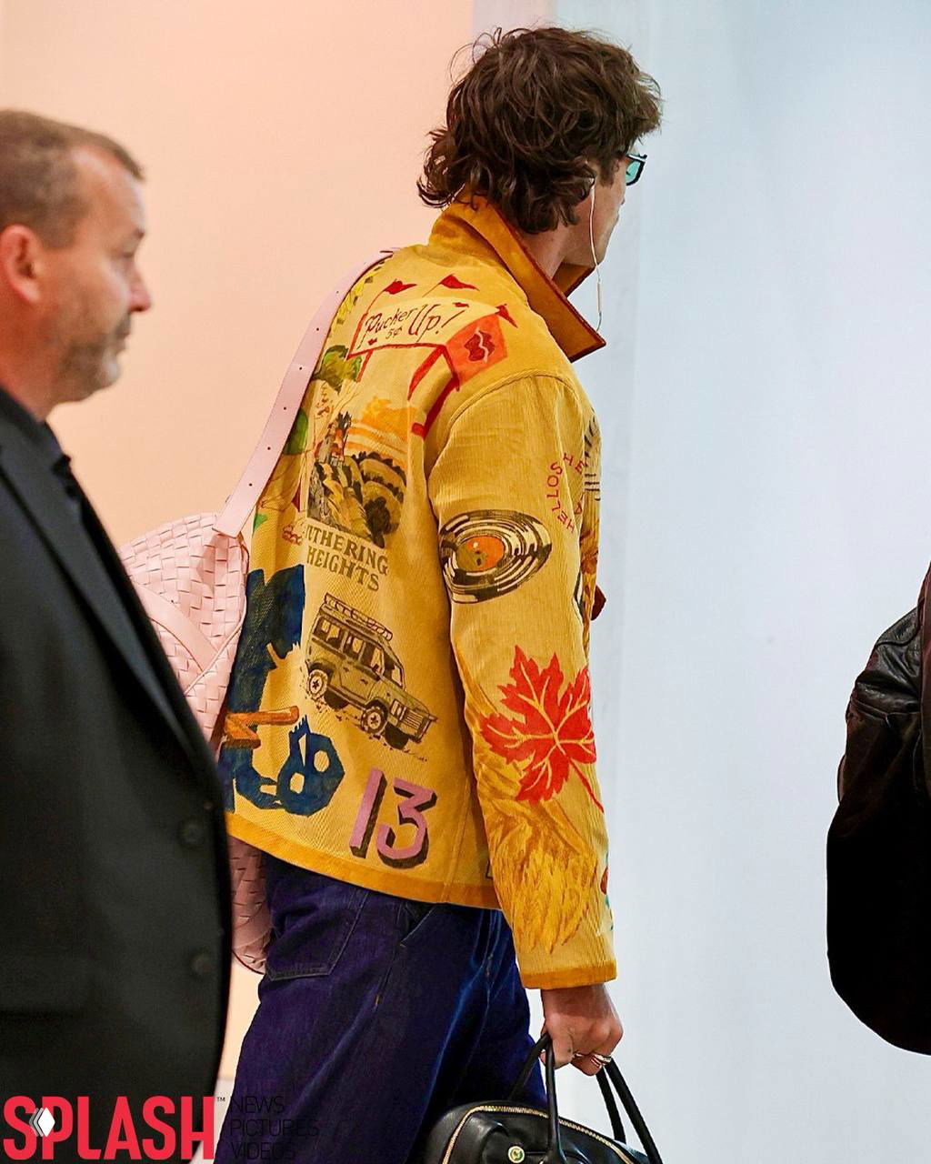 Вид со спины на мужчину в жёлтой куртке с принтами: заметны детали куртки, розовая сумка через плечо и белые проводные наушники в ушах