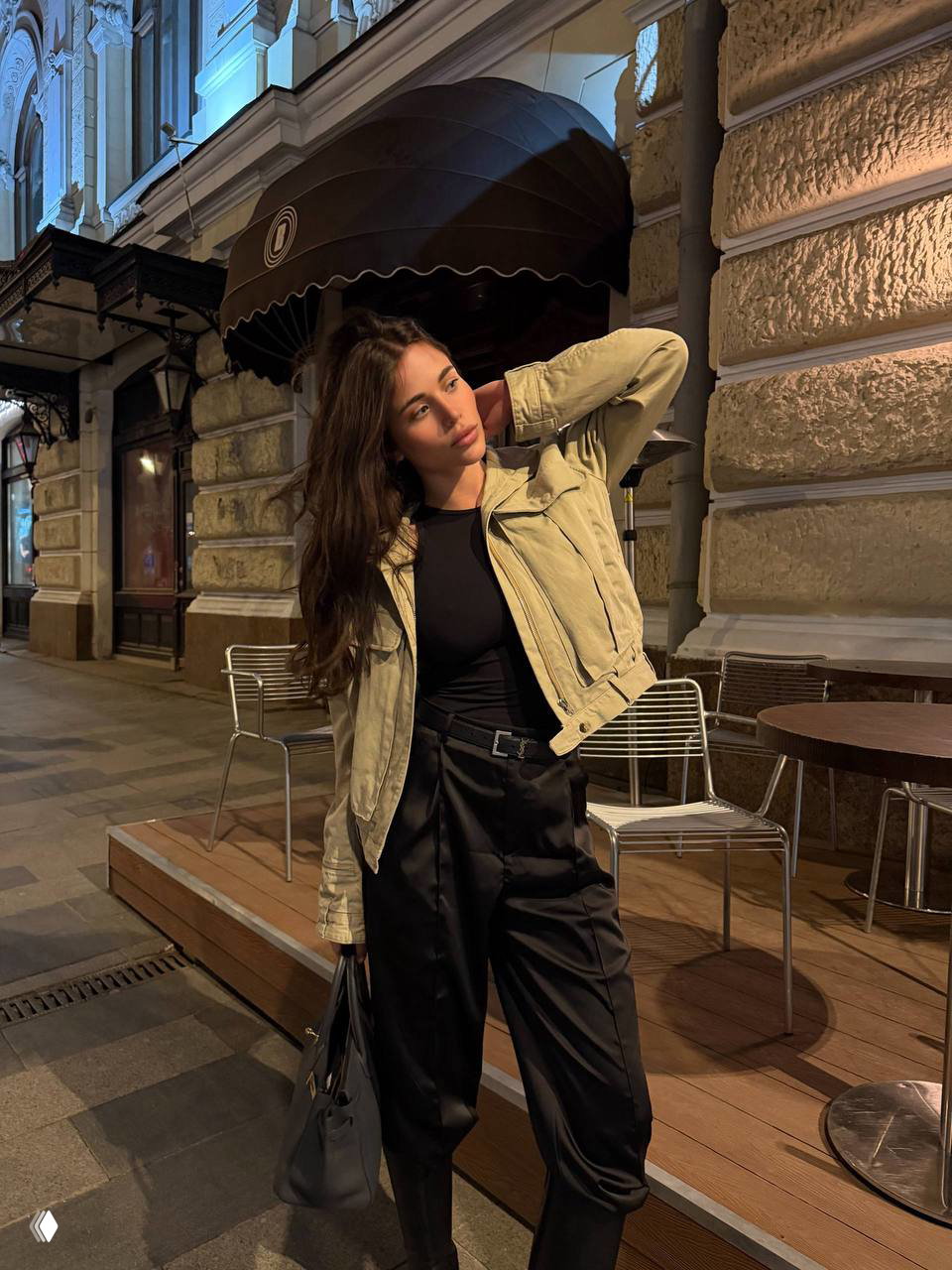 Ночная уличная портретная фотография: молодая женщина в светлой куртке и чёрных брюках позирует у фасада здания рядом с уличным кафе и креслами.