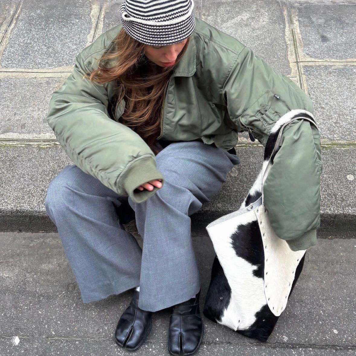 Девушка в бини и оверсайз-куртке сидит на тротуаре рядом с сумкой в пятнистом принте, напоминающем окрас далматинца — стритстайл-кадр. Фото в стиле эстетики: нейтральная палитра и мягкое освещение.