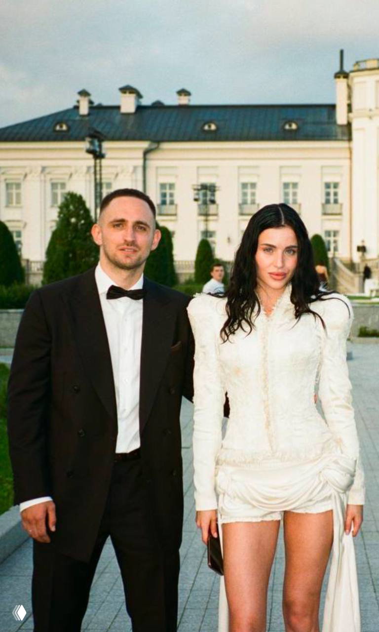 Фотография со свадебного мероприятия: пара в вечерней одежде стоит на открытом пространстве на фоне светлого исторического здания и газона позади.